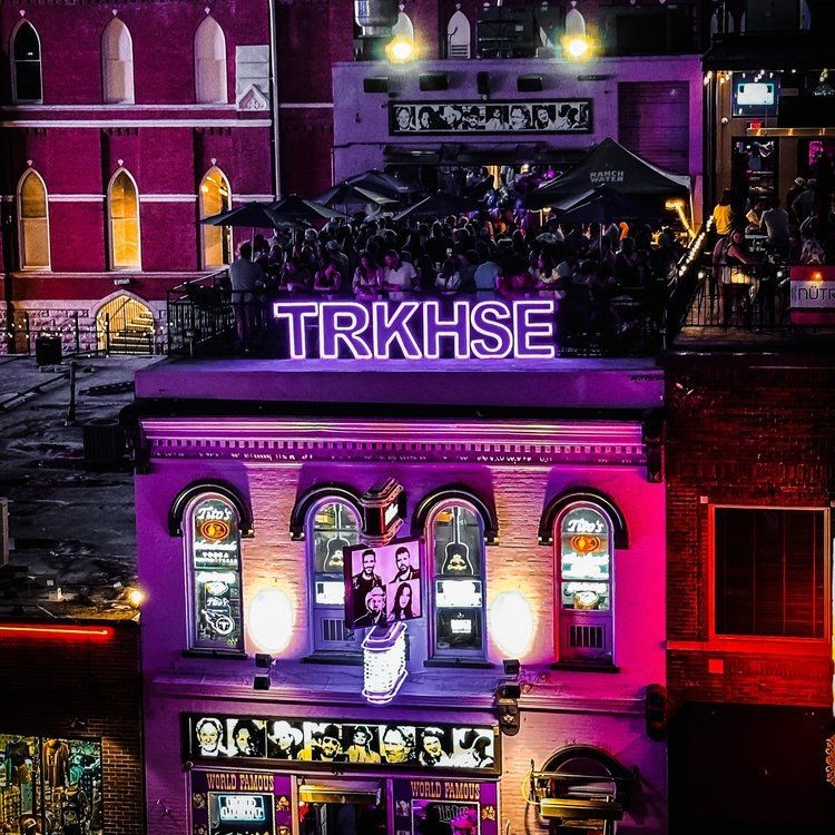 Colorful bar exterior with neon sign 'TRKHSE' and rooftop crowd at night.